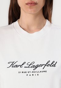 White cotton sweatshirt featuring black embroidered text: "Karl Lagerfeld 21 RUE ST-GUILLAUME PARIS." Round neckline and short sleeves.