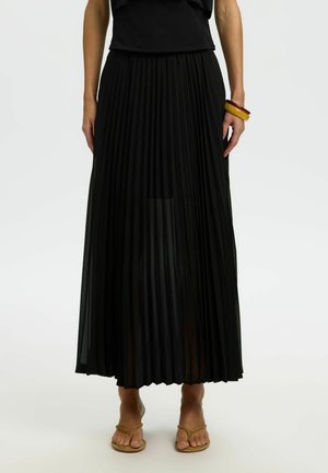 Kvinne iført svart plissett midi-skjørt, svart topp, brune sandaler og to chunky armbånd på høyre håndledd, stående på hvit bakgrunn.