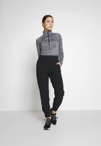 Camiseta de manga larga, gris, con cremallera, combinada con pantalones joggers negros de cintura elástica. Los pantalones tienen puños en los tobillos. Modelo que lleva zapatillas deportivas negras.