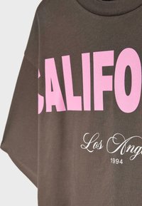T-shirt marron avec un grand texte rose "CALIFORNIA" et un texte blanc plus petit "Los Angeles 1994" en dessous à l'avant.