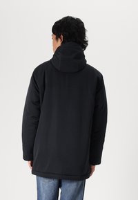 Black padded jacket with a hood, featuring a smooth texture, long sleeves, and a slightly longer back hem. Worn over light blue jeans.