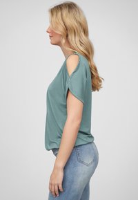 Haut à manches courtes, couleur bleu clair, avec un design à épaules dénudées, texture douce, coupe décontractée, associé à un jeans en denim bleu clair.