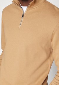 Felpa zip-up color beige realizzata in materiale testurizzato; presenta un collo alto e una zip argentata, maniche aderenti e un design casual.