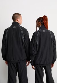 Veste coupe-vent noire avec col haut, poignets élastiques et coupe ample. Design en relief subtil et accents réfléchissants sur les épaules et le dos.