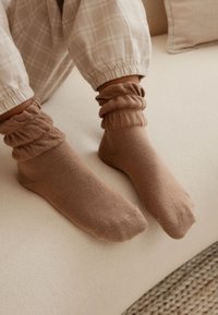 Bruine, geribbelde enkelsokken met een zachte textuur, gedragen met lichtgekleurde geruite broek, tegen een neutrale, textuurrijke achtergrond.