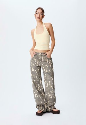 Young woman wearing a light yellow halter top, loose-fitting camouflage pants, and black platform sandals standing with hands in pockets.