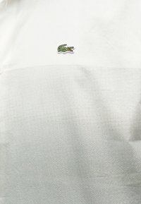 Polo blanc et gris clair avec un petit logo crocodile vert brodé centré sur la poitrine.