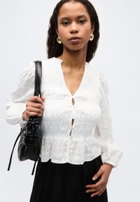 Γυναίκα με μπούκλες στα μαλλιά, που φοράει χρυσούς κρίκους στα αυτιά και ένα λευκό πουκάμισο με κουμπιά, κρατώντας μία μαύρη τσάντα ώμου, μπροστά σε ένα απλό φόντο.