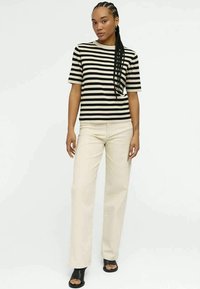 Woman wearing a black and beige striped short-sleeve top, beige wide-leg pants, and black open-toe heeled sandals, standing against a white background.