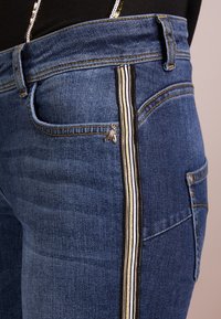 Jeans de mezclilla oscura con corte slim, franjas laterales en negro, blanco y dorado, y un pequeño emblema de abeja decorativo cerca del bolsillo delantero.