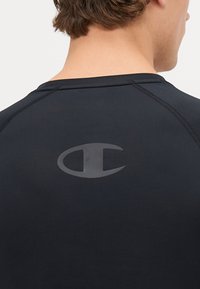 T-shirt de sport noir à manches courtes, avec une texture lisse et un logo imprimé au dos à l'envers. Détails de couture le long des épaules.