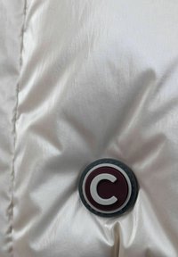 Tessuto di giacca puffy di colore chiaro con finitura lucida, caratterizzata da un pulsante logo circolare in bordeaux e bianco. Texture morbida, motivo trapuntato.