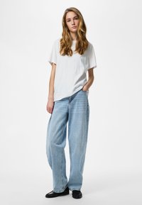 Tânără cu părul lung și blond, stând în picioare, purtând un tricou alb, blugi largi de culoare albastru deschis și pantofi negri, cu o mână în buzunar.