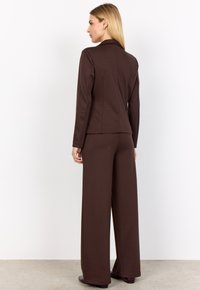 Woman with blonde hair wearing a fitted dark brown blazer and wide-leg matching trousers, standing facing away against a white background.