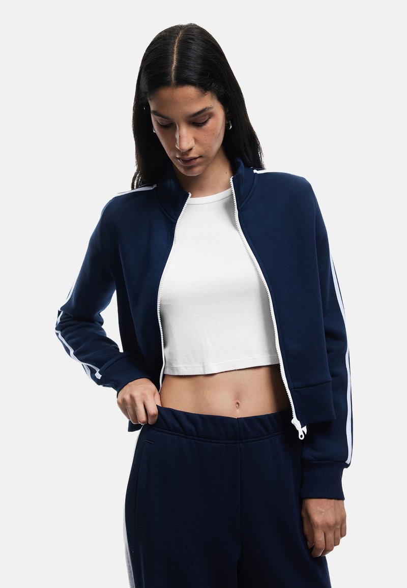 Chaqueta de cremallera azul marino con rayas blancas en las mangas, cuenta con un cuello y puños acanalados. Usada sobre un top corto blanco y joggers a juego.