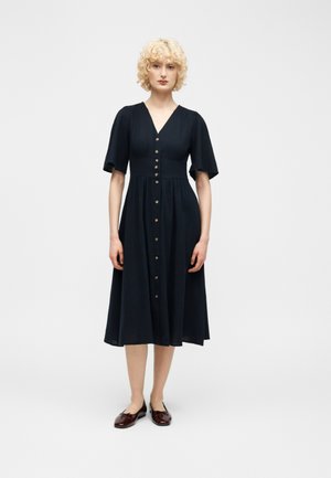 Femme blonde portant une robe noire boutonnée au genou avec des manches courtes et des chaussures plates marron foncé, debout sur un fond blanc.