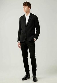 Sort dress med slank passform, enkeltspent jakke med to frontlommer, kombinert med hvit skjorte og svarte polerte sko.