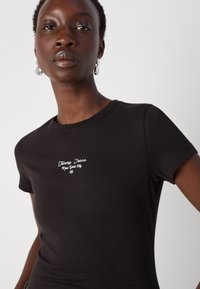T-shirt in cotone nero con maniche corte, caratterizzato da testo bianco "Tommy Jeans New York City" in corsivo, scollo rotondo e design aderente.