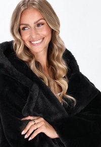 Černý kabát z umělé kožešiny s velkou kapucí, měkká textura a nadměrný design; ruka modelky ozdobená zlatými prsteny, vystavující přírodní nehty.