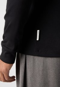 Schwarzes Langarmshirt mit einem Seitentag, kombiniert mit einer grauen Textilunterlage. Fokus auf schlankem Design und Stoffdetails.