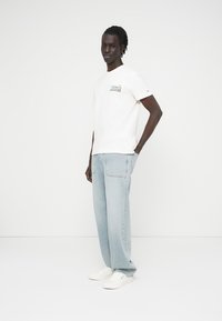 Νέος άνδρας που στέκεται με το ένα χέρι στην τσέπη ενός ανοιχτό μπλε τζιν, φορώντας λευκό T-shirt Tommy Jeans και λευκά αθλητικά παπούτσια σε ουδέτερο φόντο.