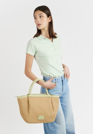 Giovane donna con polo verde chiaro e jeans blu che tiene in mano una borsa intrecciata beige con bordatura verde chiaro su sfondo bianco.