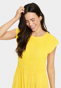 Yellow dress featuring a smocked bodice and short sleeves, with soft fabric and flowing skirt. The texture is gathered with a neat finish.