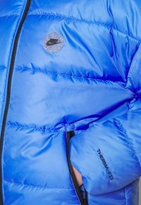 Niebieska kurtka puchowa wykonana z błyszczącego materiału, z okrągłym logo Nike, zamkami błyskawicznymi i napisem "THERMA-FIT" na rękawie, zapewniającym izolację.