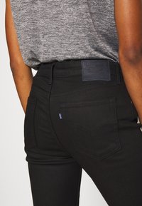 Jeans de mezclilla negra con cintura alta, bolsillos traseros y una etiqueta de parche de cuero negro en la cintura con detalles de costura azul.