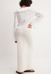 Weißes Langarm-Bluse mit rüschigem Ausschnitt, kombiniert mit einem langen, strukturierten, gehäkelten Rock im Rautenmuster. Schwarze Flip-Flops.