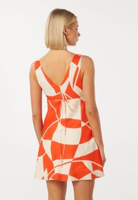 Kurzes, ärmelloses Kleid mit V-Ausschnitt auf dem Rücken. Es hat auffällige geometrische Muster in Orange und Creme und ist an der Taille mit einem Bindeband gerafft.