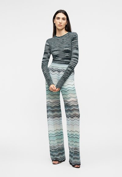 Žena oblečená v pruhovanom top s dlhými rukávmi a vysokými širokými nohavicami so vzorom cikcak, stojaca so zloženými rukami.