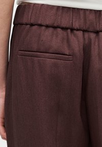 Κοντινό πλάνο σκούρων καφέ παντελονιών με ελαστική μέση και μια μονή τσέπη τύπου welt στο πίσω μέρος, φορεμένα με λευκό τοπ.