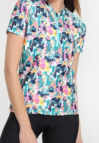 Mujer con un top deportivo de manga corta, colorido con flores y cremallera, y pantalones cortos de ciclismo ajustados negros, torso visible sobre un fondo liso.