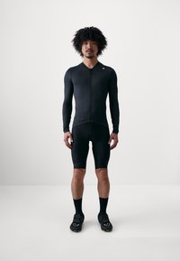 Svart sykkeldrakt med kort glidelås foran, lange ermer og tettsittende shorts. Materialet ser glatt ut med subtile merkevare detaljer.