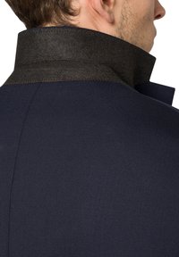 Ciemnoniema płaszcz z wysokim kołnierzem, wykonanym z kontrastującego, teksturowanego ciemnobrązowego materiału. Widoczne szwy wzdłuż krawędzi kołnierza.