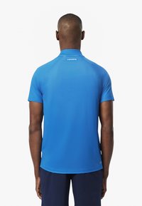 Sininen lyhythihainen polo-paita, jossa on sileä ja kevyt kangas. Paidassa on kaulus ja merkin "LACOSTE" painatus selkäpuolella kaula-aukkoa lähellä.