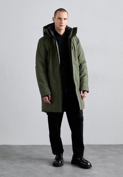Olive green insulated parka with a hood, front zipper, and two side pockets. Worn over a black top and black pants, featuring smooth texture.