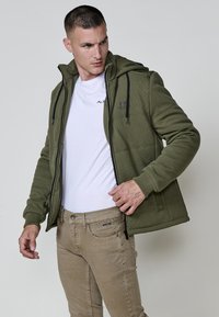 Tamsiai žalia zip-up striukė su tekstūruotu dizainu, gobtuvu ir juodais virvutėmis. Dėvima ant baltos T-shirt ir derinama su šviesiai rudos spalvos kelnėmis.