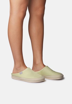 Inimene, kes kannab helerohelisi libisesussid beežide talladega, seisab tavalise valge tausta ees.