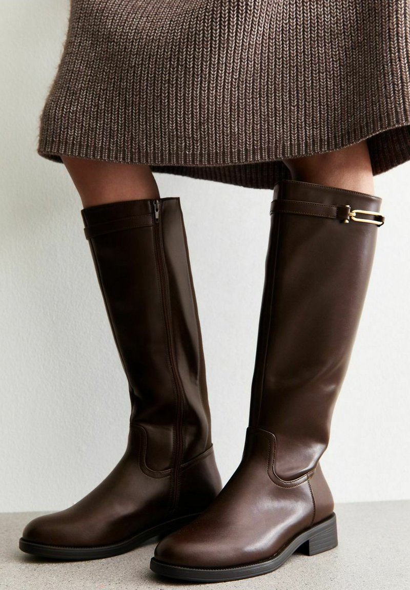 Knee-high dark brown leather boots with side zipper and gold buckle detail worn with a brown knitted skirt.