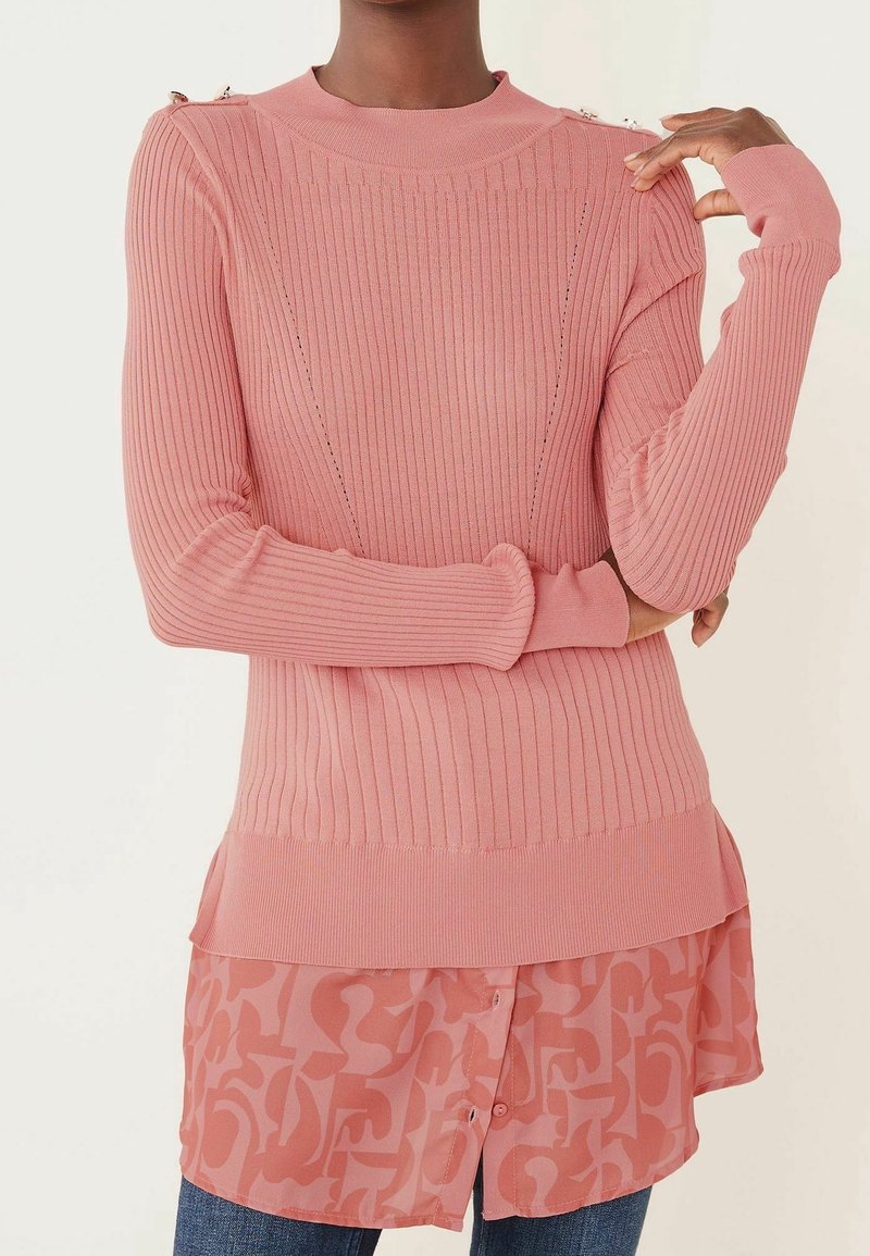 Vrouw in een geribbelde roze trui met knopen op de schouders, gedragen over een gemusterde roze blouse en blauwe jeans.