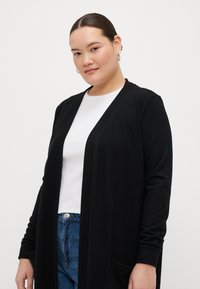 Ilgas juodas kardiganas pagamintas iš minkštos medžiagos, turi atvirą priekį, dvi priekines kišenes ir ilgas rankoves, derinamas su baltu marškinėliu ir mėlynomis džinsinėmis kelnėmis.