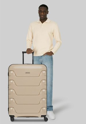 Mann in beigem Langarmhemd und Jeans steht mit beigem Hartschalenkoffer mit vier Rädern und ausgezogenem Griff vor grauem Hintergrund.