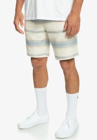 Pruhované plážové šortky v krémové, modré a pískové barvě. Vyrobené z texturované látky s uvolněným střihem, spárované s bílými ponožkami a teniskami.