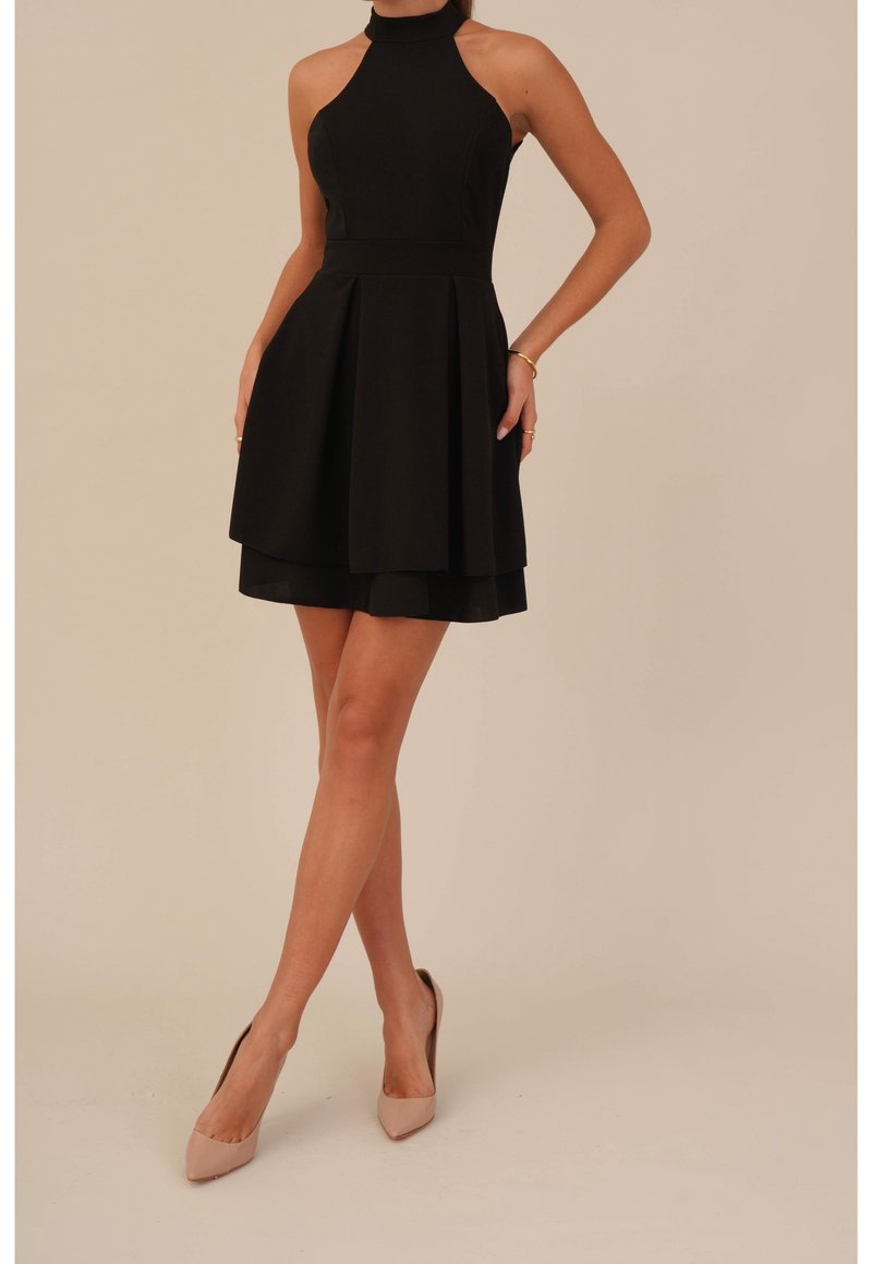 Femme portant une robe noire sans manches à col halter avec une jupe évasée et des talons hauts beige, debout, jambes croisées et mains sur les hanches.