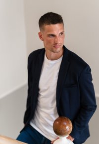 Mand iført en marineblå blazer over en hvid t-shirt, der står nær et træ- og hvidt trappegelænder med et rundt træaccent.