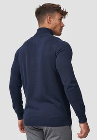Mørkeblå strikket genser med høy krage, ribbestrikkede mansjetter og kant, tettsittende design og glatt tekstur, båret av en modell sett fra ryggen.