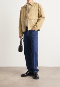 Beżowa kurtka z kanwy z zamkiem błyskawicznym, dwoma przednimi kieszeniami, w połączeniu z ciemnoniebieskimi dżinsami i czarnymi butami. W ręku trzymana jest mała czarna torba.