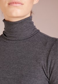 Nahaufnahme einer Person, die einen dunkelgrauen gerippten Rollkragenpullover trägt, wobei der Hals, das Kinn und die oberen Schultern zu sehen sind.